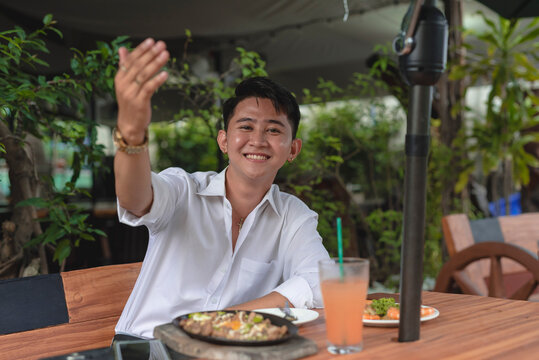 An Asian young guy orders a meal for two and does a "come here" hand gesture as he invites his friend over for lunch.
