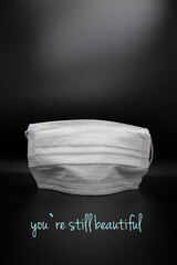 white medical mask with black background in covid 19 times with inscription you are still beautiful                    