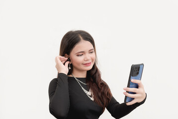 A studio shot of a cute young woman taking a selfie using her brand new cellphone while holding her bluetooth earbuds. Isolated on a plain white background.