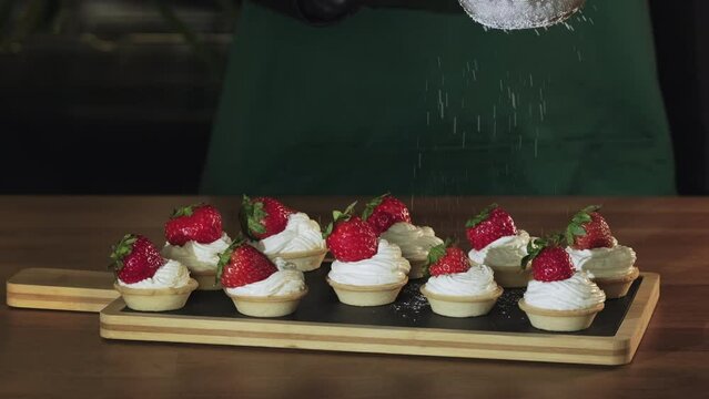 Chef sprinkling sugar powder on tasty mini cake with cream and strawberries, slow motion. Powdered sugar being sieved on strawberry tart. Dessert dusted with castor sugar