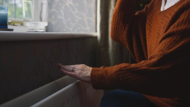 Close up of senior woman sitting by radiator at home trying to keep warm during energy and cost of living crisis drinking hot drink and shivering - shot in slow motion 