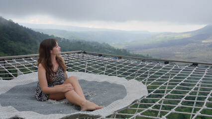 Beautiful woman sitting on a hammock net couch outdoors above green misty abyss. Korean female tourist chilling on a rug high in the mountains with a splendid view on valley and volcano. Happy holiday