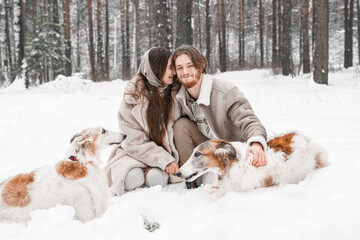 Love romantic young couple girl, guy in snowy cold winter forest walking with pet, dog of hunting breed russian borzoi. Sighthound, wolfhound owner. Having fun, laughing. Stylish fur coat, woolen hat
