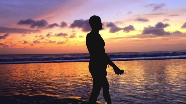 Silhouette of a man walking along the ocean against the backdrop of a bright fiery multi-colored sunset. A man enjoying the peace and sound of the waves. Slow motion 4k footage of beautiful landscape.