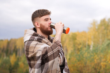 Portrait of handsome guy, young happy man is enjoy drinking tea or coffee, hot beverage from thermo cup, thermos bottle in plaid, cozy blanket in natural golden autumn park or forest, smile - obrazy, fototapety, plakaty