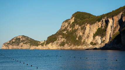 Rocky cliffs with some trees at Liapades bay lit by morning sun, calm sea water and boat buoys in foreground