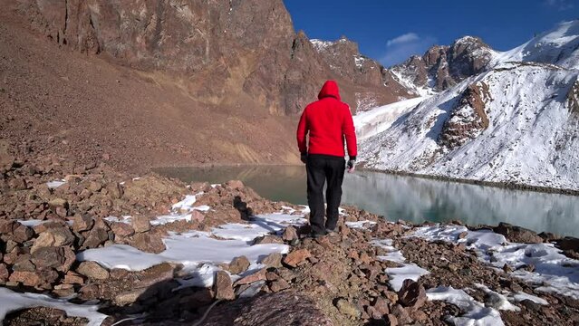 A guy in a tracksuit goes to a moraine lake in the mountains. An ancient snow glacier among high rocks. The guy is wearing gloves, a red jacket and black pants. Blue sky with clouds. Almaty Mountains