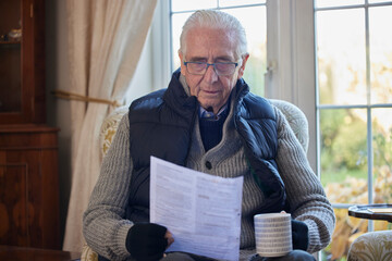 Senior Man Wearing Extra Clothes With Hot Drink Trying To Keep Warm At Home In Energy Crisis...