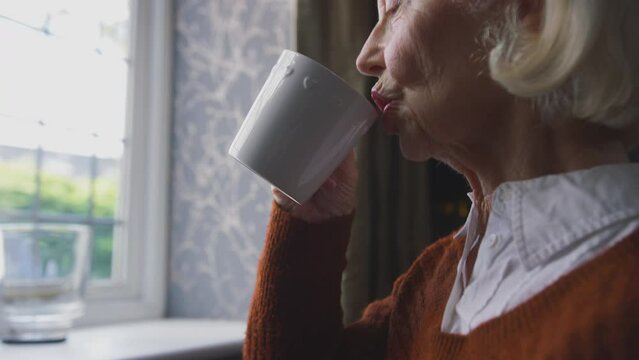Senior woman sitting by radiator at home trying to keep warm during energy and cost of living crisis drinking hot drink and shivering - shot in slow motion 