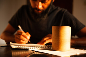 brown skinned person with a grown beard writing on a notebook and a brown coffee mug on a side taking notes