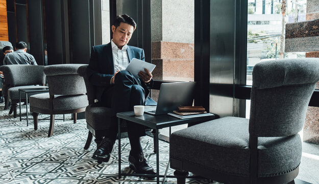 Handsome Asian man in elegant blue suit reading business report on papers and writing notes while sitting in the cafe with a laptop computer and cup of coffee