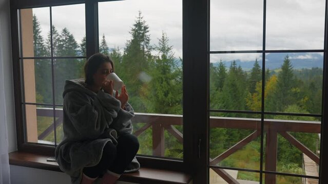 Woman holding mug, sitting on windowsill and looking at mountains