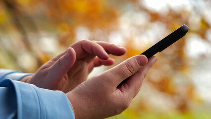 Female hands in light blue sleeves work on smartphone. Woman touches and swipes screen of mobile phone on autumn day on blurred background closeup