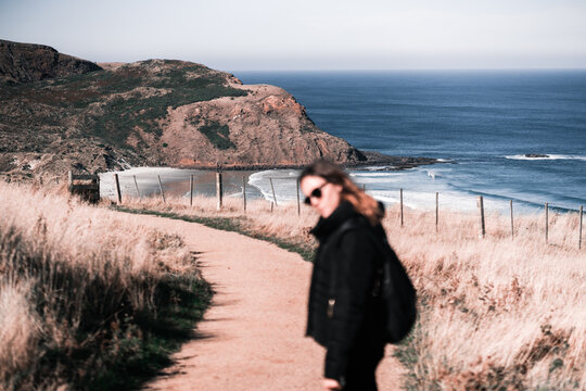 caucasian girl in black jacket and sunglasses looking at camera standing on path leading to calm beautiful beach on sunny day, sandfly bay, new zealand