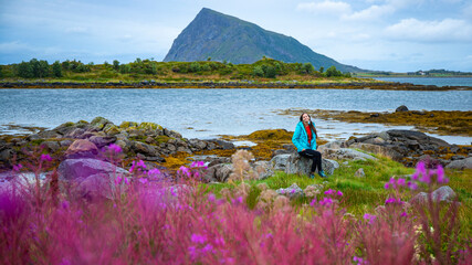 A beautiful girl sits on moss surrounded by lush purple flowers with huge mountains in the background; lofoten islands, norway and its fjords