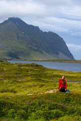 girl in red sweatshirt sits on moss under huge cliffs in the Norwegian fjords, panorama of lofoten...