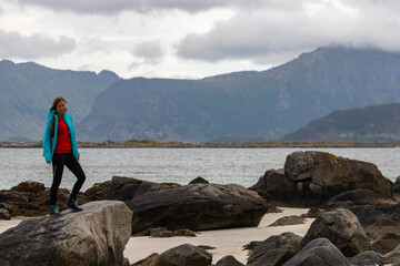 Fototapeta premium girl in blue jacket walks on the beach with huge rocks and mountains in the background, the Lofoten islands in norway and their harsh landscapes