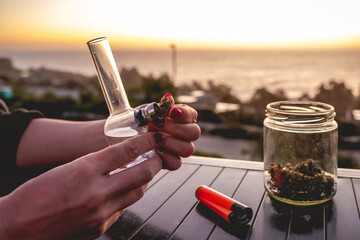 Hands with red nails putting a weed bud in a marijuana bong (weed water pipe), glass jar with marijuana buds and orange lighter with beautiful sunset sky and sea