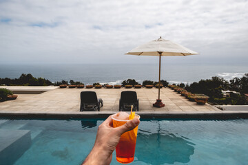 Hand holding a glass with orange alcohol cocktail in front of a big pool in terrace with chairs and parasol and the ocean in a summer evening