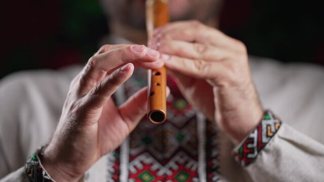 Hands of man playing on woodwind wooden flute - ukrainian sopilka on dark background. Folk music concept. Musical instrument. Musician in traditional embroidered shirt - Vyshyvanka.