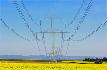 Strommast mit Kabeln und Isolatoren vor blauem, wolkenlosen Himmel mit blühendem Rapsfeld