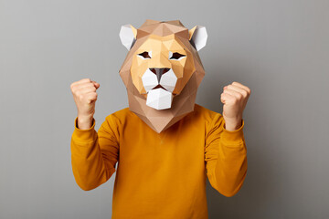 Portrait of unknown man wearing lion mask and orange jumper isolated over gray background, standing...