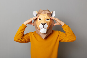 Portrait of unhealthy anonymous man wearing lion mask and orange sweatshirt isolated over gray background, having headache, keeps hand on head, suffering pain, being tired.