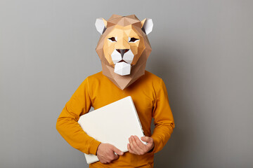 Obraz premium Indoor shot of unknown man wearing lion mask and orange jumper isolated over gray background, standing holding closed notebook in hands, looking at camera.