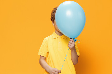 a boy of school age stands on an orange background in a yellow polo and covers his face with a large blue balloon