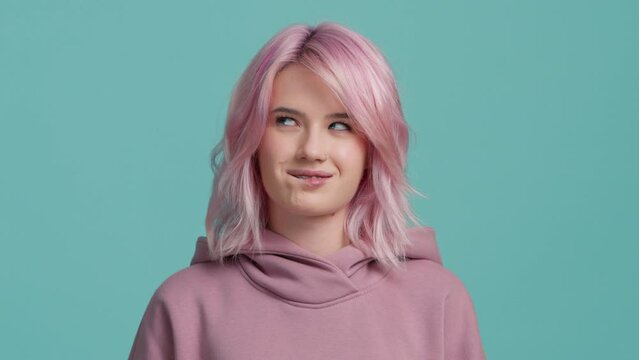 Cunning young woman with pink color hairstyle in hoodie thinking devious tricks cheats, sly squint, bites her lower lip and scheming evil plan, trying to plot something sly in mind on blue background