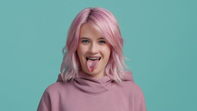 Cheerful funny hipster student girl with pastel pink hair showing tongue with piercing, smiling with brackets on white teeth, making faces at camera, and laughing fooling around. Pretty female studio