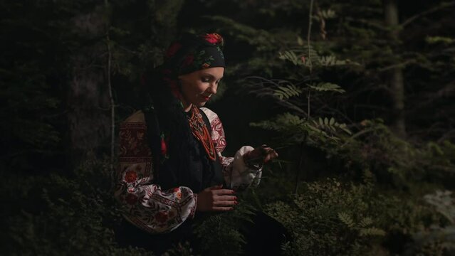 Witch woman collects herbs, ferns at night in Carpathian mountains forest. She in in traditional ukrainian handkerchief, national dress - vyshyvanka, ancient coral beads. Folk medicine concept.