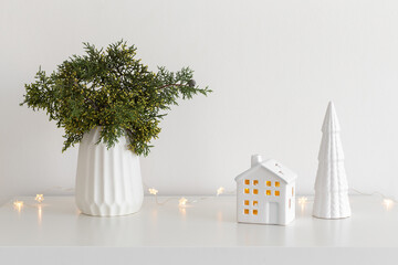 Christmas cozy winter home decor. New year interior decorations. Green fir branch in vase, decorative ceramic house and christmas tree, glowing garland lights. Stylish composition on the table.