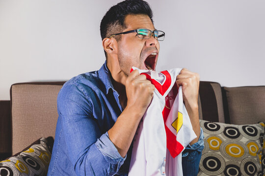 Joven fan&aacute;tico del f&uacute;tbol, gritando gol, apoyando a su equipo sentado en el sof&aacute; de casa viendo la transmisi&oacute;n en vivo del encuentro. Concepto de mundial de futbol e hinchas del futbol en el mundo.