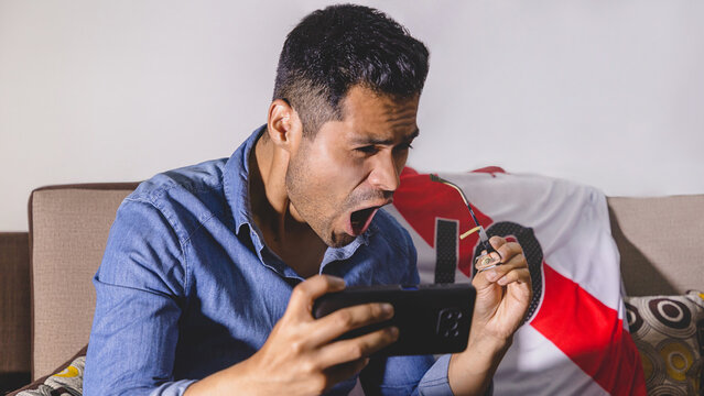 Joven peruano hincha del f&uacute;tbol, selecci&oacute;n peruana de futbol. Gritando gol, apoyando a su equipo sentado en el sof&aacute; de casa viendo la transmisi&oacute;n en vivo. Concepto de mundial de futbol e hinchas del f