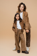 cheerful woman holding hand with daughter posing in trendy autumn outfit on beige background