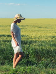Naklejka na ściany i meble Young woman in white dress and hat walking in the wheat field on a sunny spring day