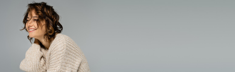 young woman with wavy hair smiling in knitted cardigan isolated on grey, banner