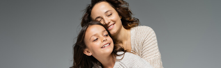 trendy woman smiling with closed eyes near happy daughter isolated on grey, banner