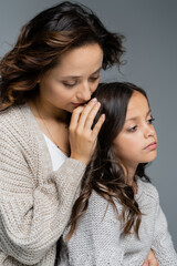 young woman in knitwear hugging head of brunette daughter isolated on grey
