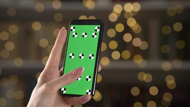 Woman hand holds vertically phone green screen and her finger scroll zoom on background bokeh blur. Close up mockup phone with markers for using tracking and replacing video on display. Use online map