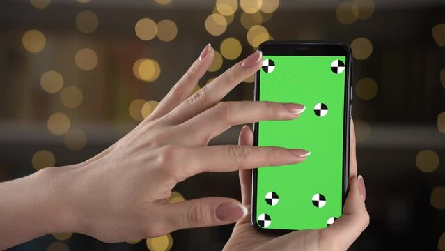 Woman using vertically mock up phone swipes his finger across the green screen and zooms map. In hands gadget with markers for tracking and inserting images on display. Blurred background bokeh.
