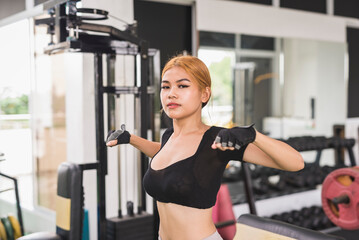 Fototapeta na wymiar An asian woman in a black crop top looks at the camera while doing dynamic chest stretch before a workout session at the gym.