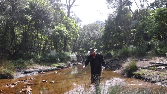 A bushman in a stockman jacket crosses a wild creek out in the Australian bush while looking for water.
