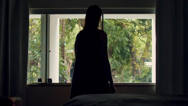 Silhouette of woman looking out bedroom window after waking up in the morning. Slow motion