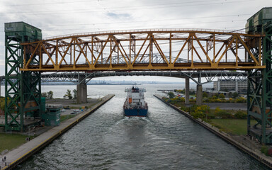 A bulk carrier cargo ship, blue and white, passes under the two large truss bridges, entering a large industrial port.