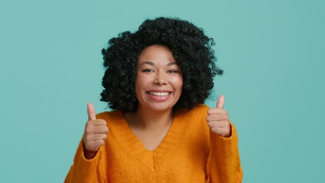 Young attractive African American woman 20s wearing casual sweater looking at camera showing thumbs up like gesture isolated against over plain teal blue background, studio portrait curly hair model