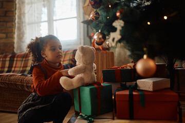 Little black girl playing with plush toy by Christmas tree at home. - obrazy, fototapety, plakaty