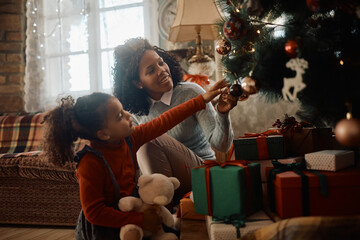 Happy African American mother decorating Christmas tree with her small daughter.