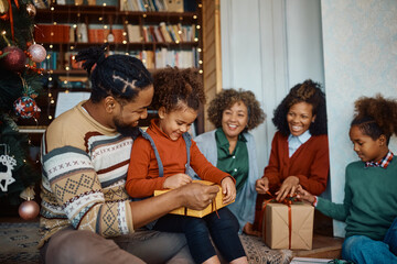Happy black little girl opens gift box while celebrating Christmas with her family at home.